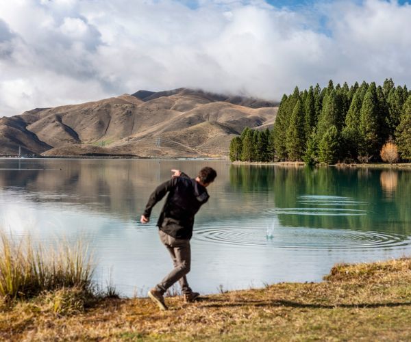 A man in a black sweater and pants throwing a pebble into a lake